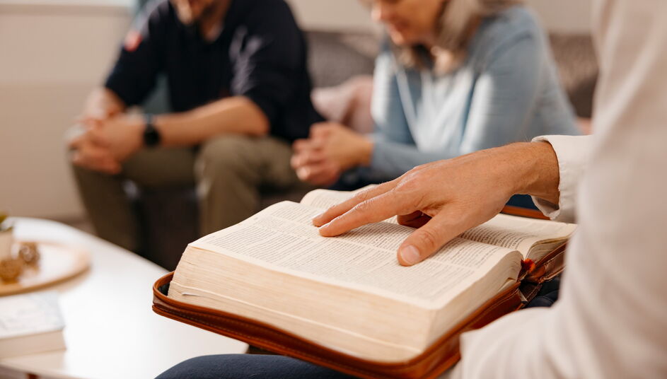 Eine Person zeigt auf eine Textstelle in einer aufgeschlagenen Bibel. Im Hintergrund sitzen zwei weitere Personen zusammen und tauschen sich aus.