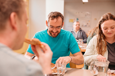 Mehrere Personen sitzen gemeinsam an einem Tisch und geniessen eine Mahlzeit. Ein Mann mit Brille und türkisem T-Shirt lächelt, während er isst.