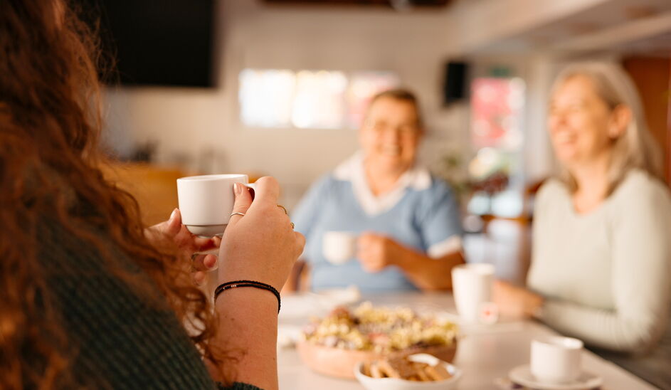 Drei Frauen sitzen gemeinsam an einem Tisch, trinken Kaffee und unterhalten sich in gemütlicher Atmosphäre. Auf dem Tisch stehen Gebäck und kleine Snacks.