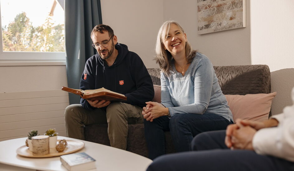 Ein Mann liest aus einer aufgeschlagenen Bibel vor, während eine Frau neben ihm lächelt. Gemeinsam sitzen sie mit einer weiteren Person in einem gemütlichen Wohnzimmer.