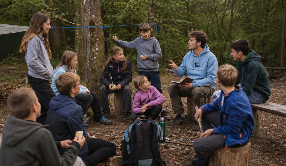 Symbolbild (KI-generiert): Eine Gruppe von Kindern und Jugendlichen sitzt mit Betreuungspersonen im Wald zusammen. Ein Leiter hält ein Buch in der Hand und spricht zur Gruppe.