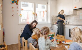 Mehrere Kinder spielen und basteln gemeinsam an einem Tisch, während sie von erwachsenen Betreuungspersonen begleitet werden.
