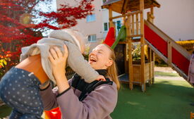 Eine lachende Betreuungsperson hebt ein Kleinkind auf dem Spielplatz in die Höhe. Im Hintergrund steht ein hölzernes Spielgerüst mit Rutsche.