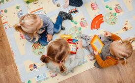 Drei Kleinkinder sitzen gemeinsam auf einem bunten Spielteppich und beschäftigen sich mit verschiedenen Spielsachen.